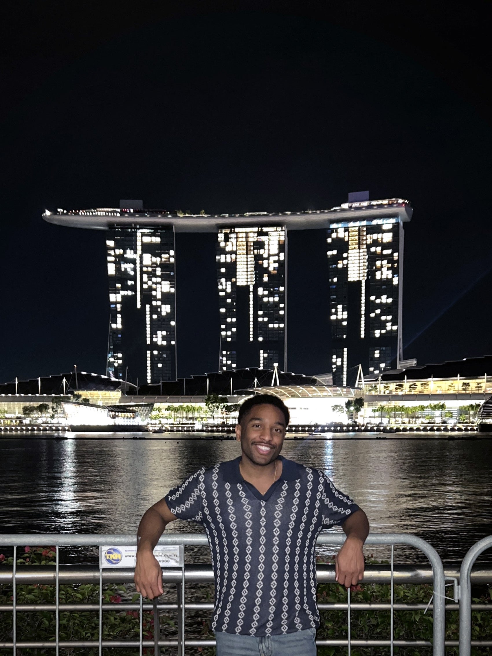 A man leans against a metal gate across the river from three tall modern buildings