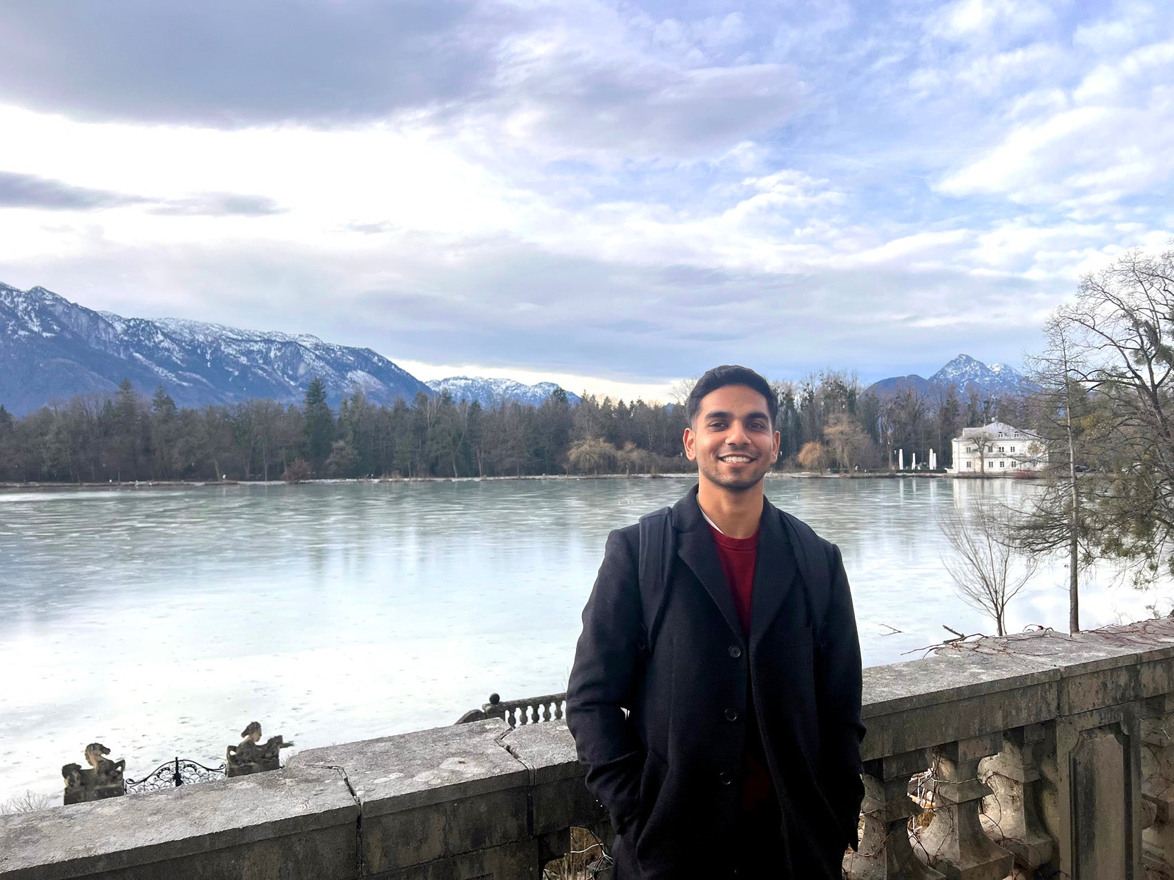 A man stands against a stone railing along the side of a river with mountains in the background.