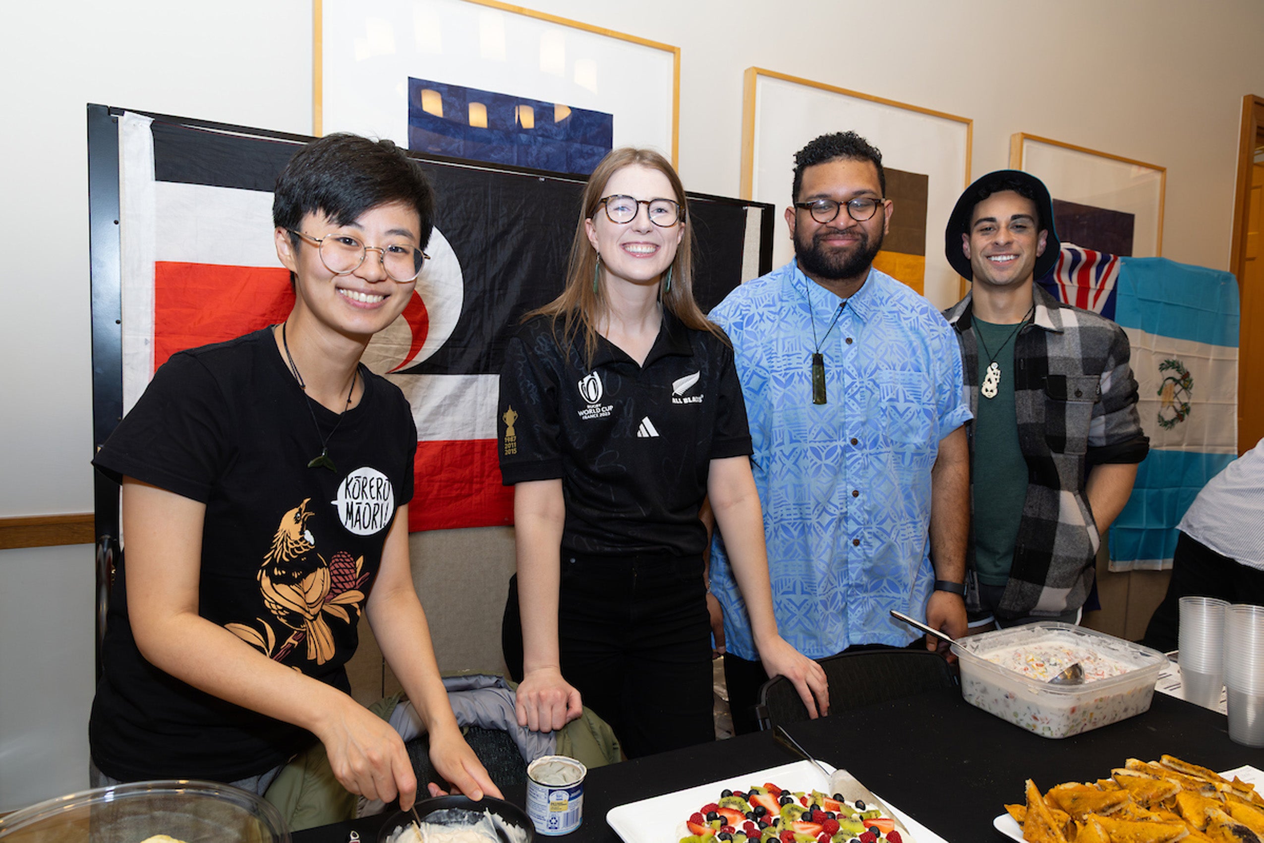 Four students stand behind a table of food in front of a flag.