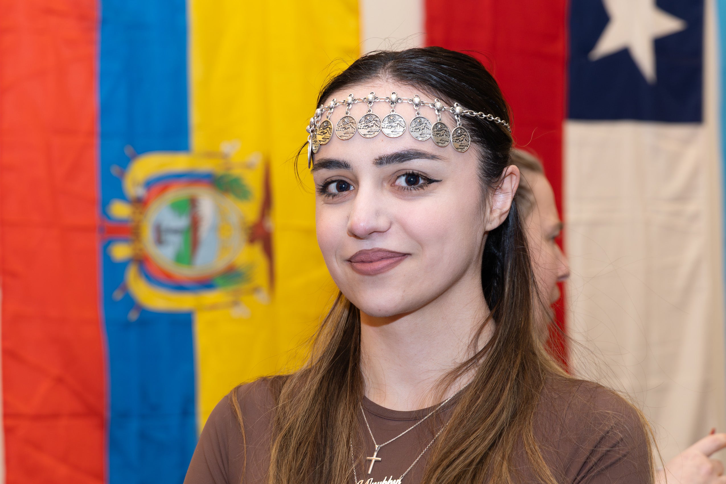 A woman wearing a decorative headpiece stands in front of a flag at an event.