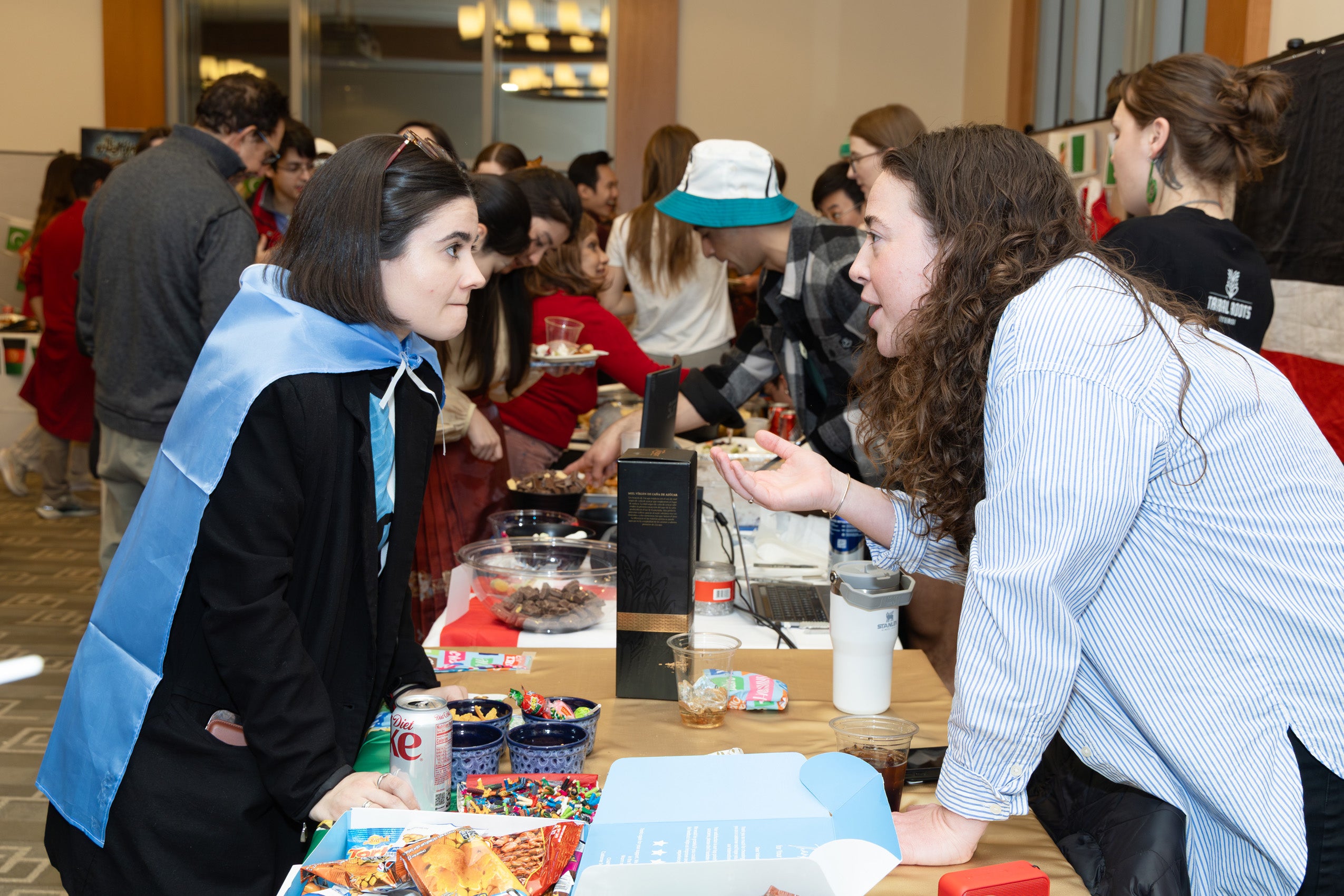 A women leans over a table to explain something to another woman at an international party.