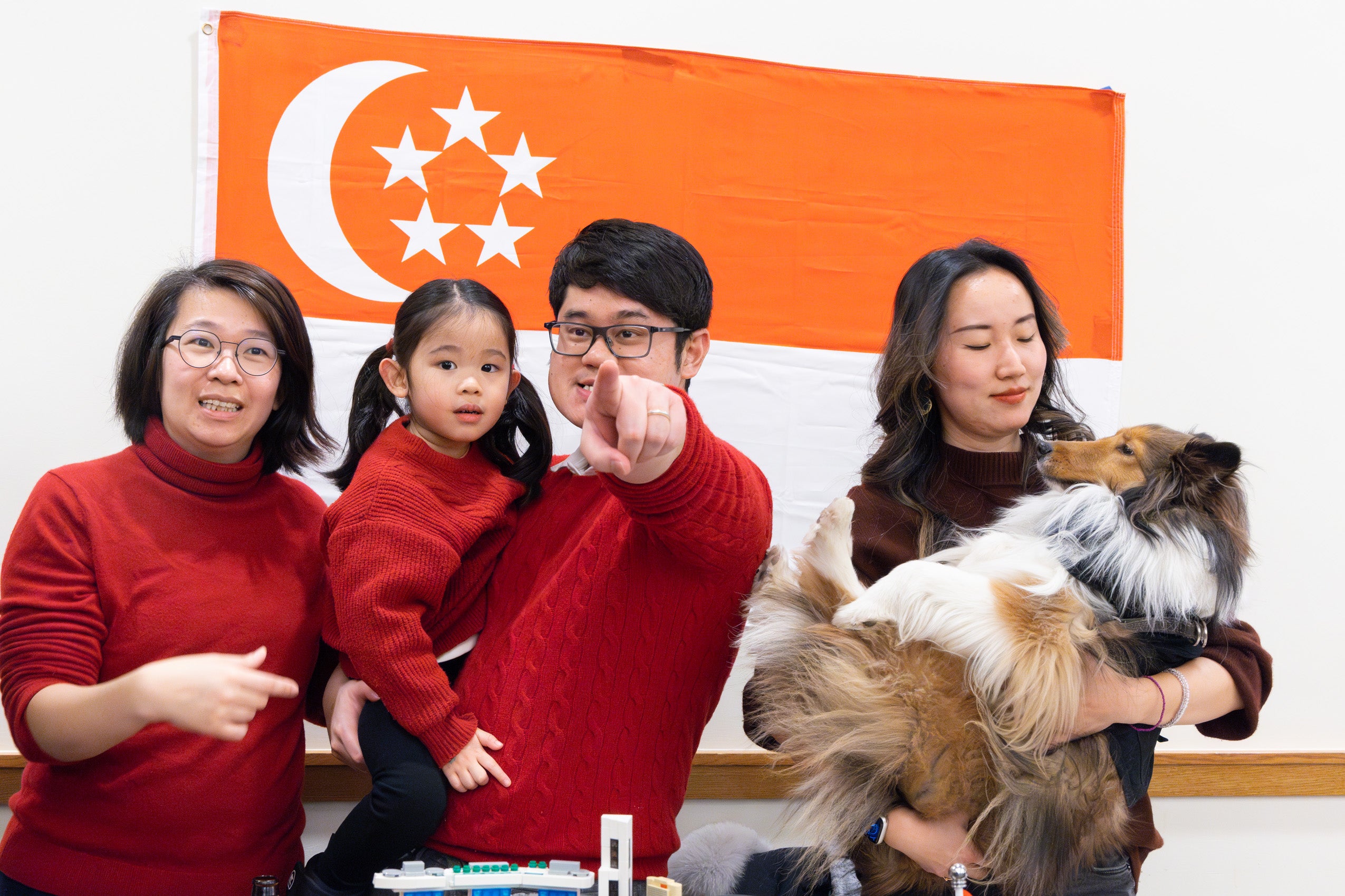 A family with a young child and a woman with a dog stand in front of a flag at an international party.