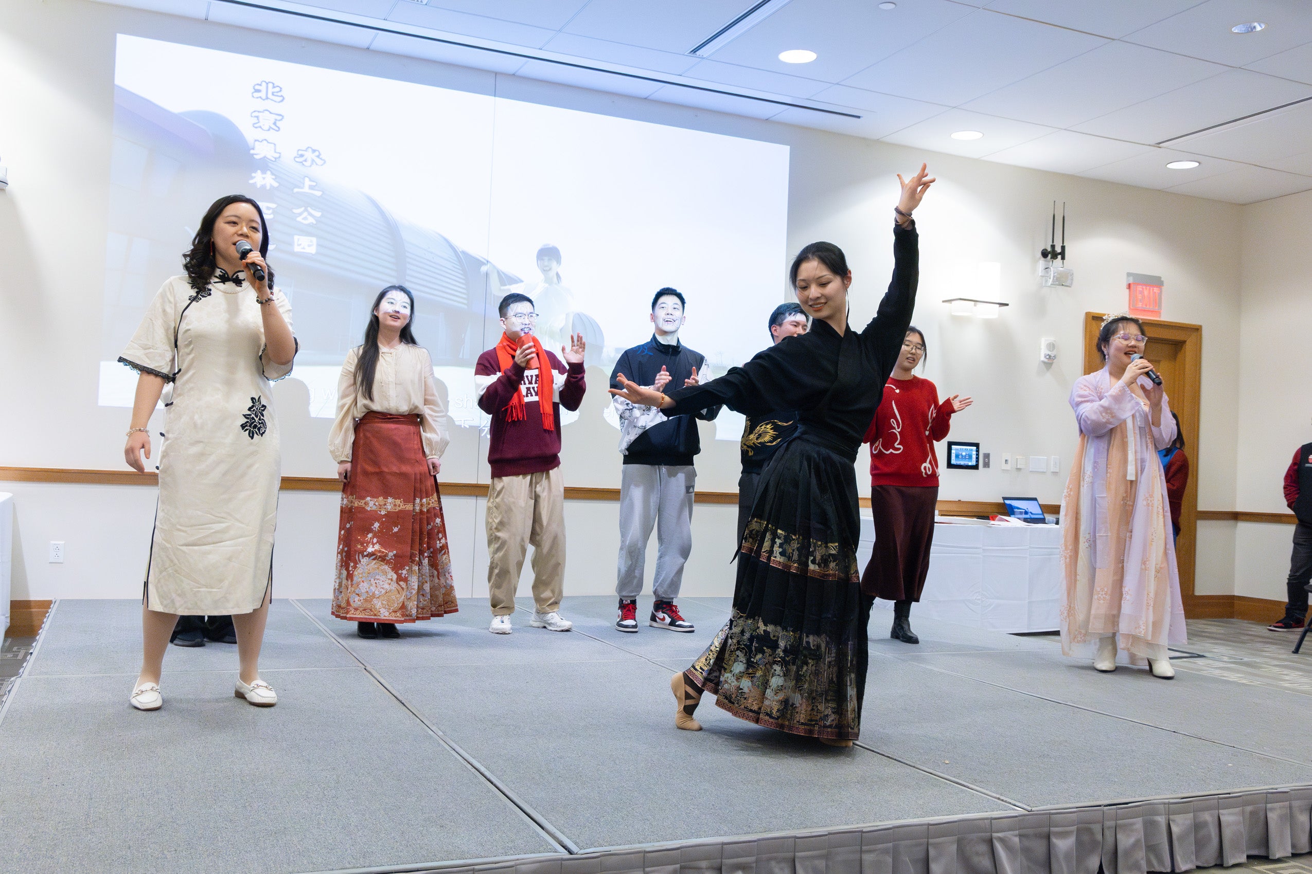 A woman in a black dress performs on stage as several others in the background sing.