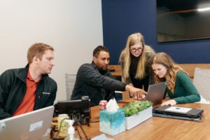 Sitting around a table in an office students point to information on a computer screen ast others look on.