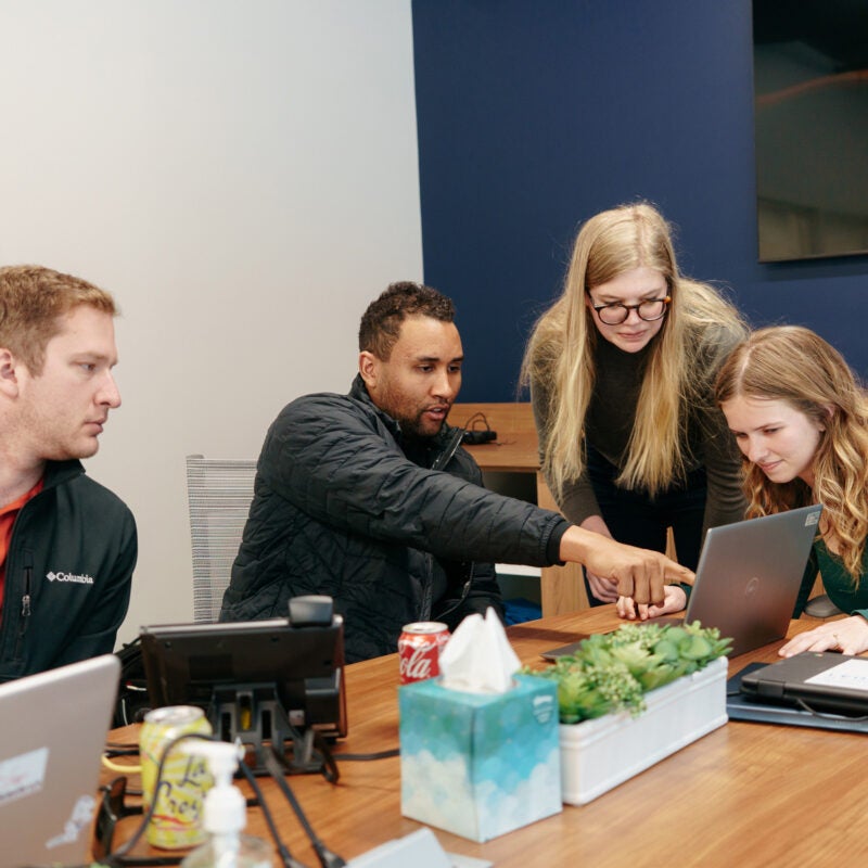 Sitting around a table in an office students point to information on a computer screen ast others look on.