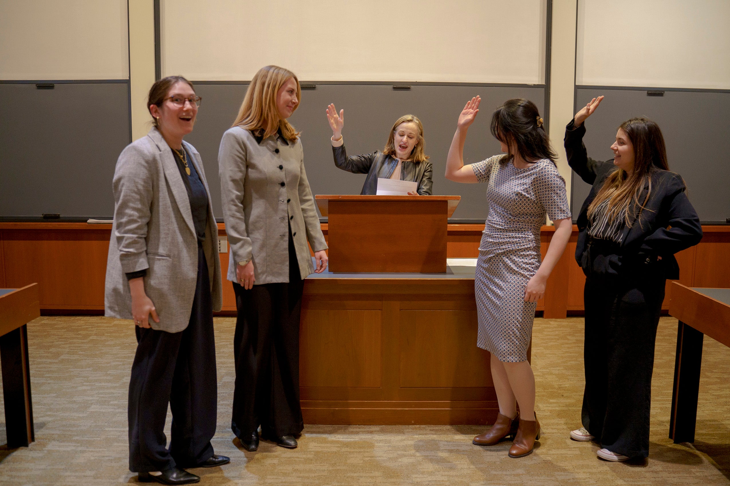 A woman makes an announcement from a podium in a classroom as others nearby respond with hi fives.