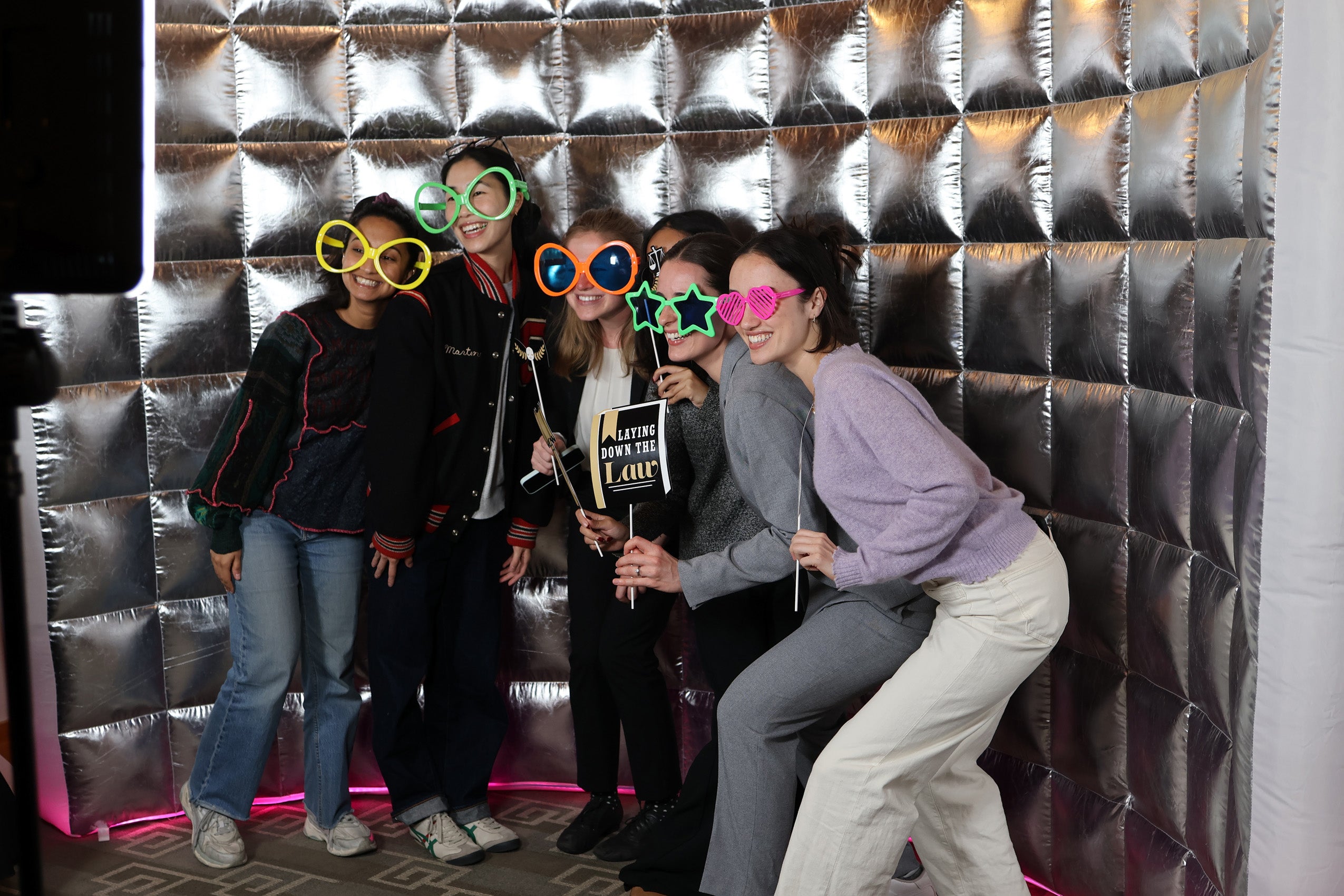 Students pose in front of a silver screen camera booth wearing colorful glasses.