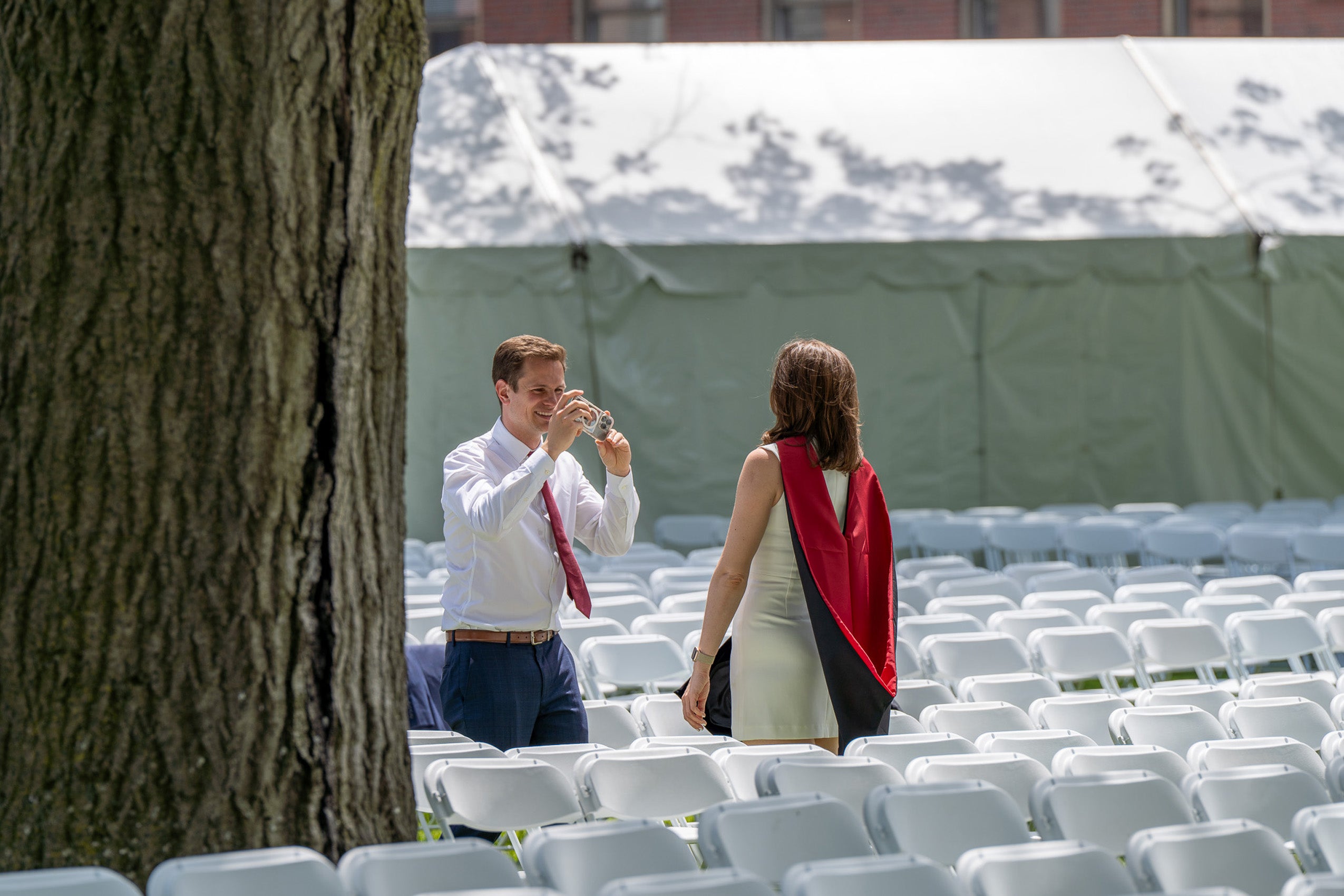 A man takes a photo of a women wearing commencement attire standing amid rows of white chairs.