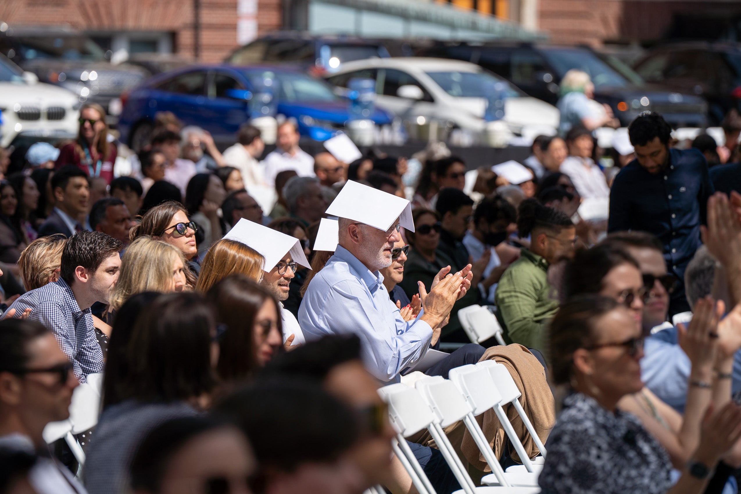 Several people sitting in seats watching class day activities wear event programs on their heads for shade.