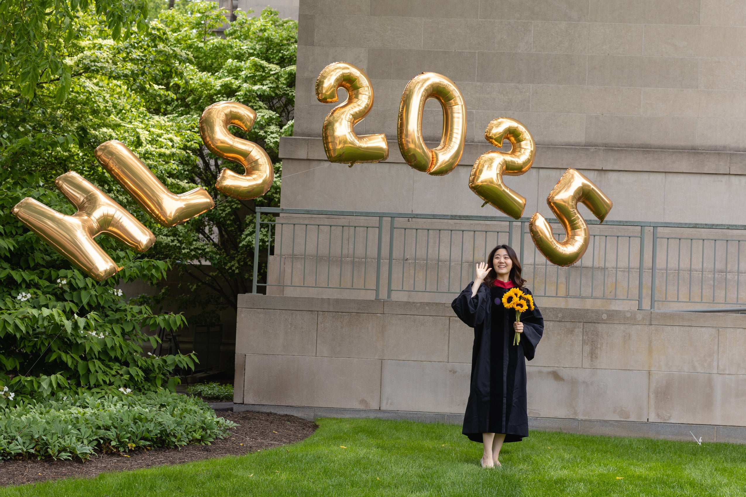 A woman holding yellow daisies poses under golden Harvard Law School balloons.