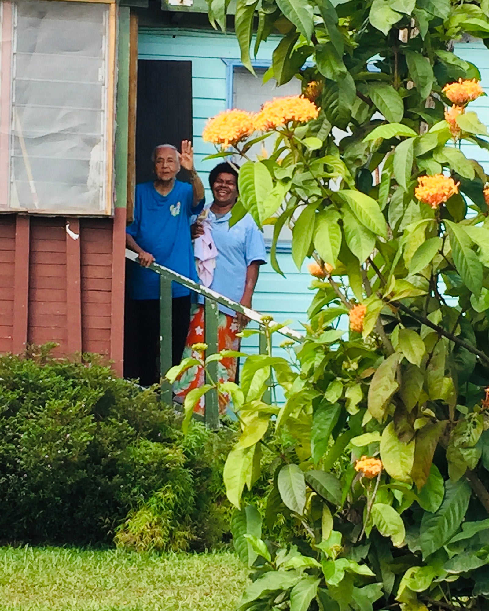 Esther Gabriel's grandmother waves from the staircase of her house with a flower tree in the foreground.