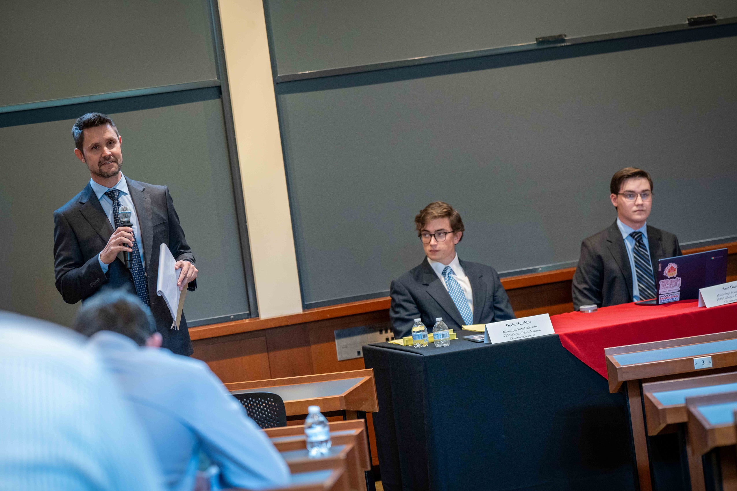 A man holding a microphone speaks next to students on a panel in a classroom.