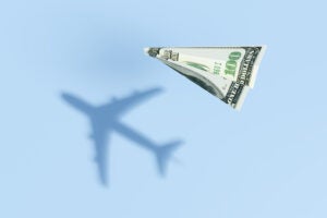Paper plane made of a $100 bill casting a shadow of a commercial flight.