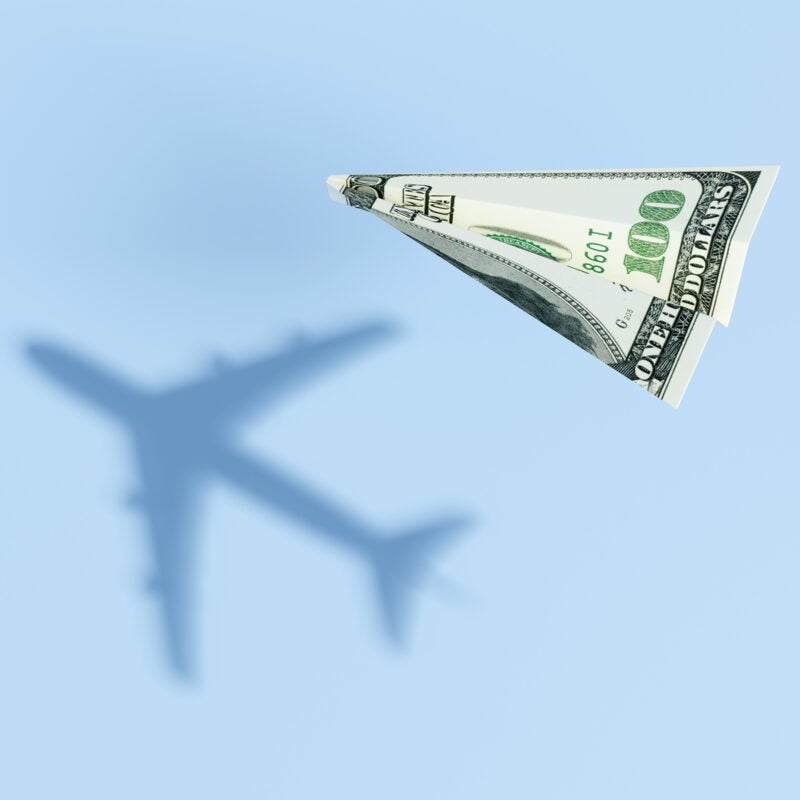 Paper plane made of a $100 bill casting a shadow of a commercial flight.