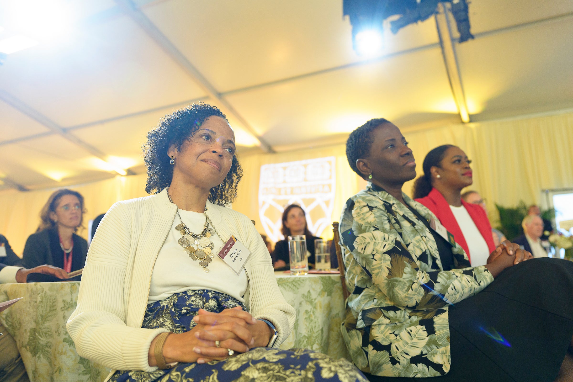 Several people listen from tables to a speaker at an event.