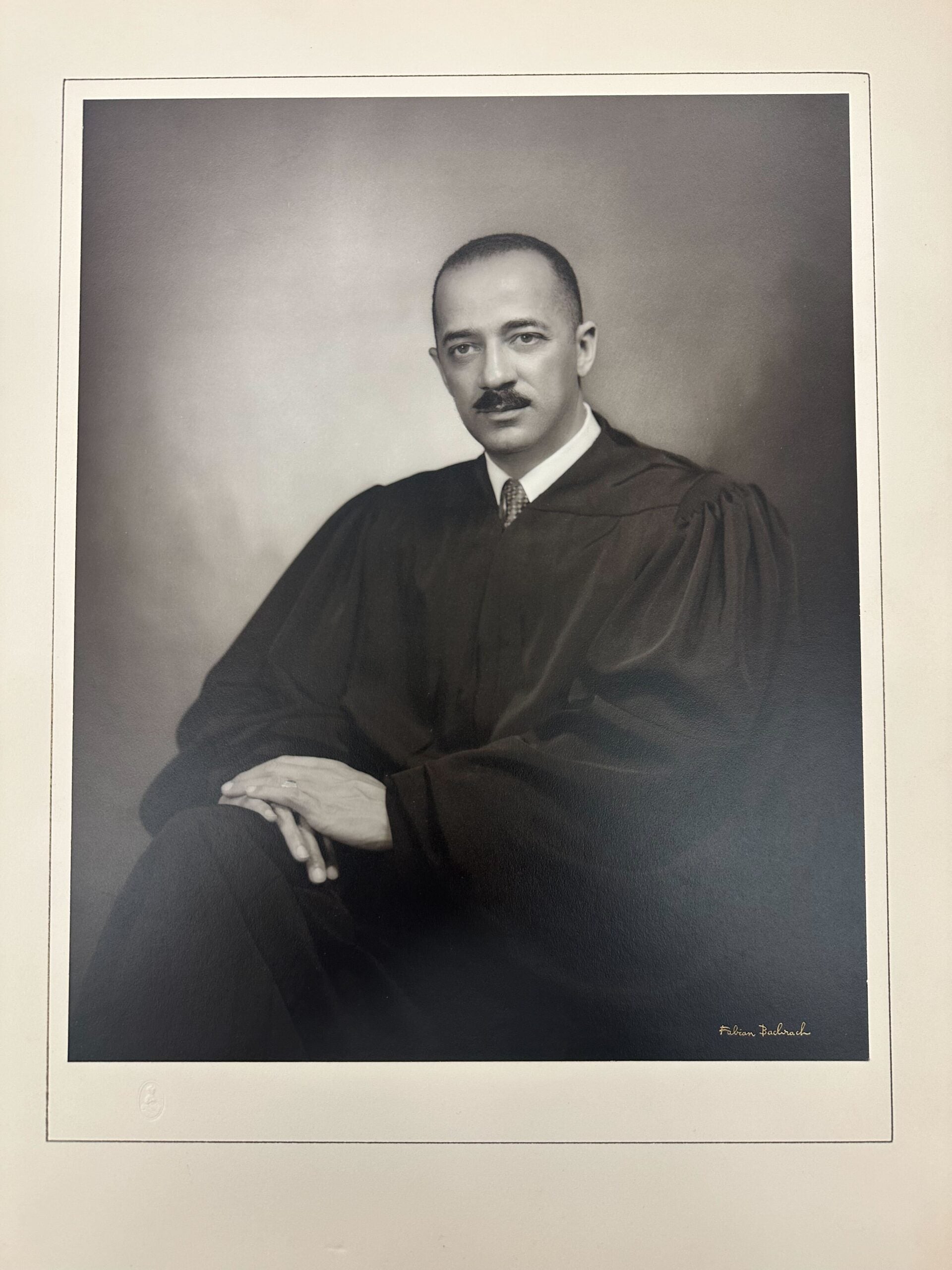 Formal posed black and white portrait of William H. Hastie in judicial robes sitting with legs crossed and hands resting on knee, looking to camera, in front of plain background, photographer’s printed signature Fabian Bachrach in bottom right corner.