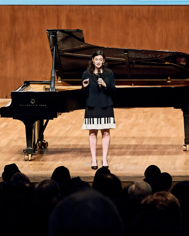 A woman holding a microphone stands in front of a piano.