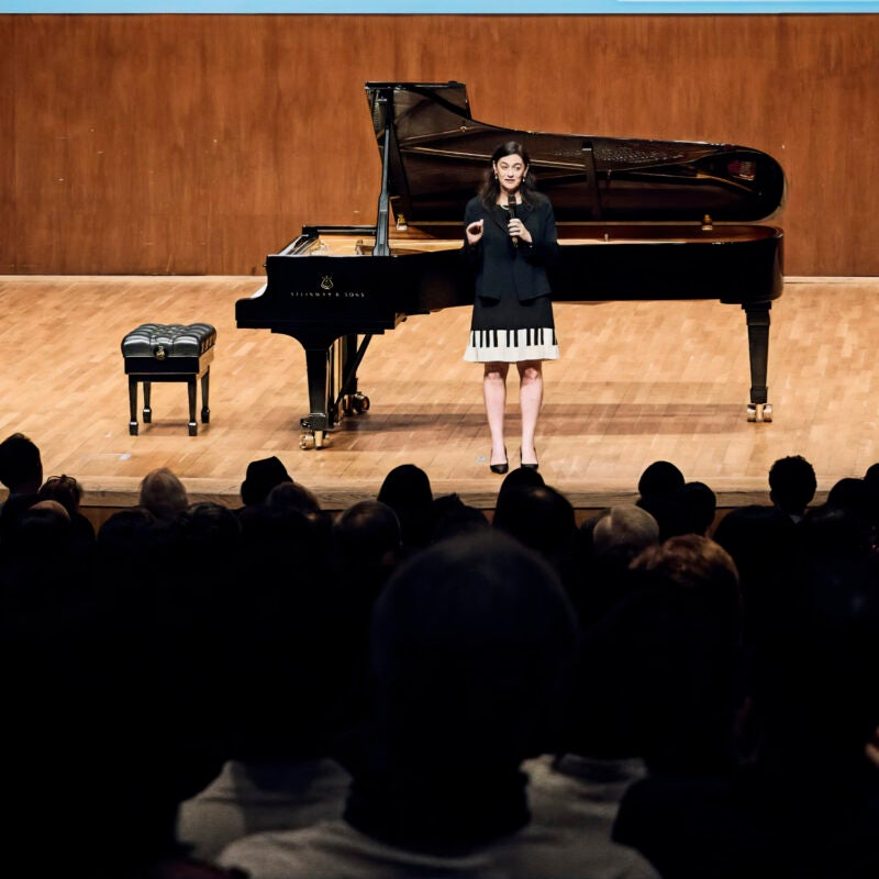 A woman holding a microphone stands in front of a piano.