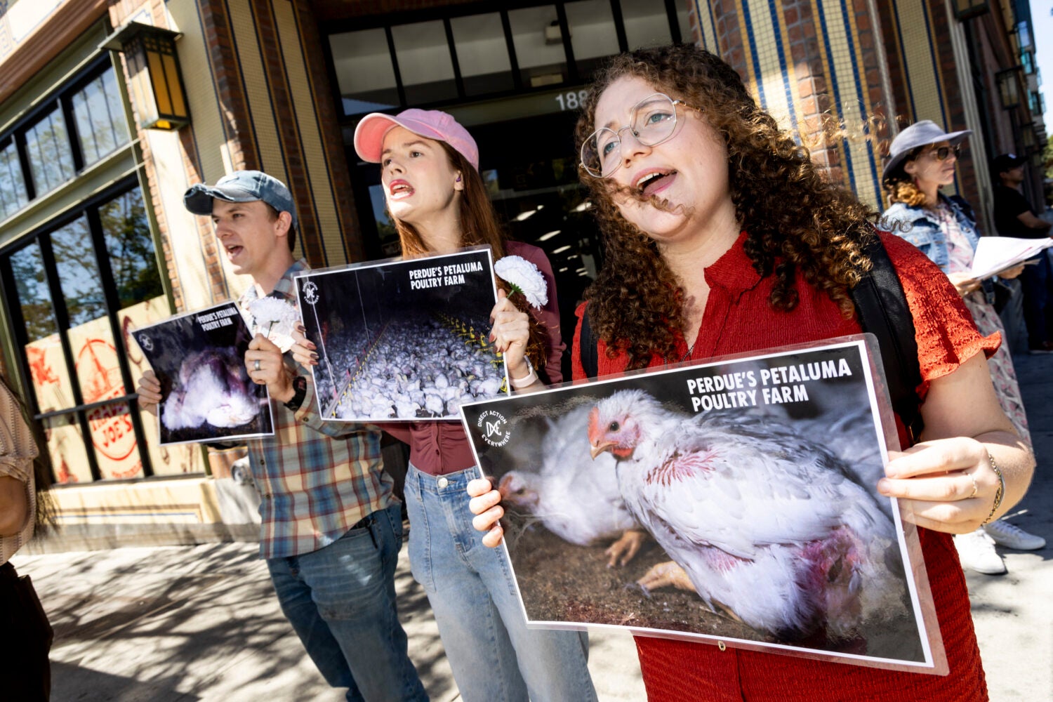 Three people stand in front of a store holding signs protesting a poultry farm.