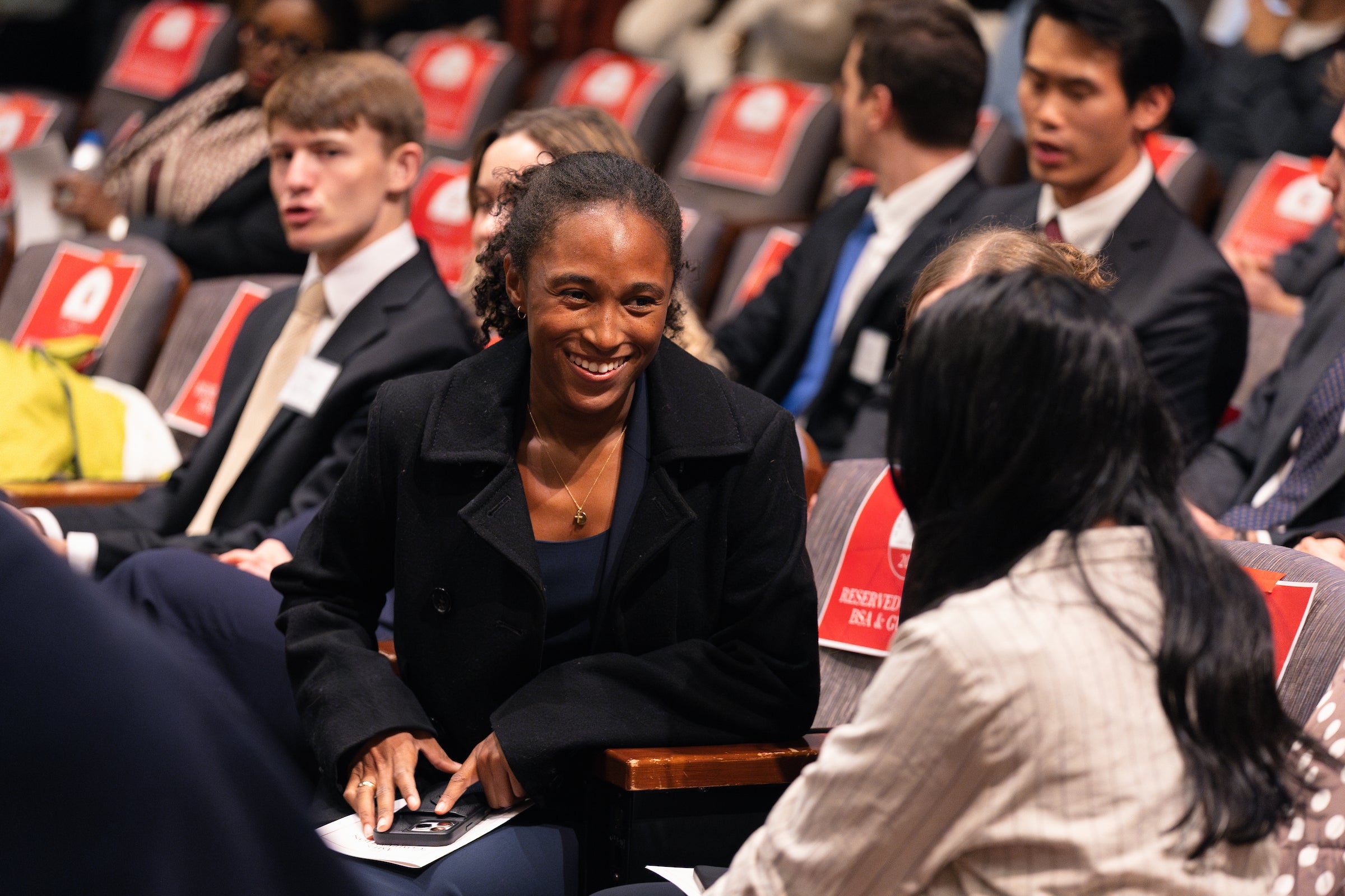 Two women sitting in an audience chat as they wait for an event to begin.