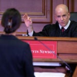 A woman stands at a podium as she speaks to a judge on the bench.