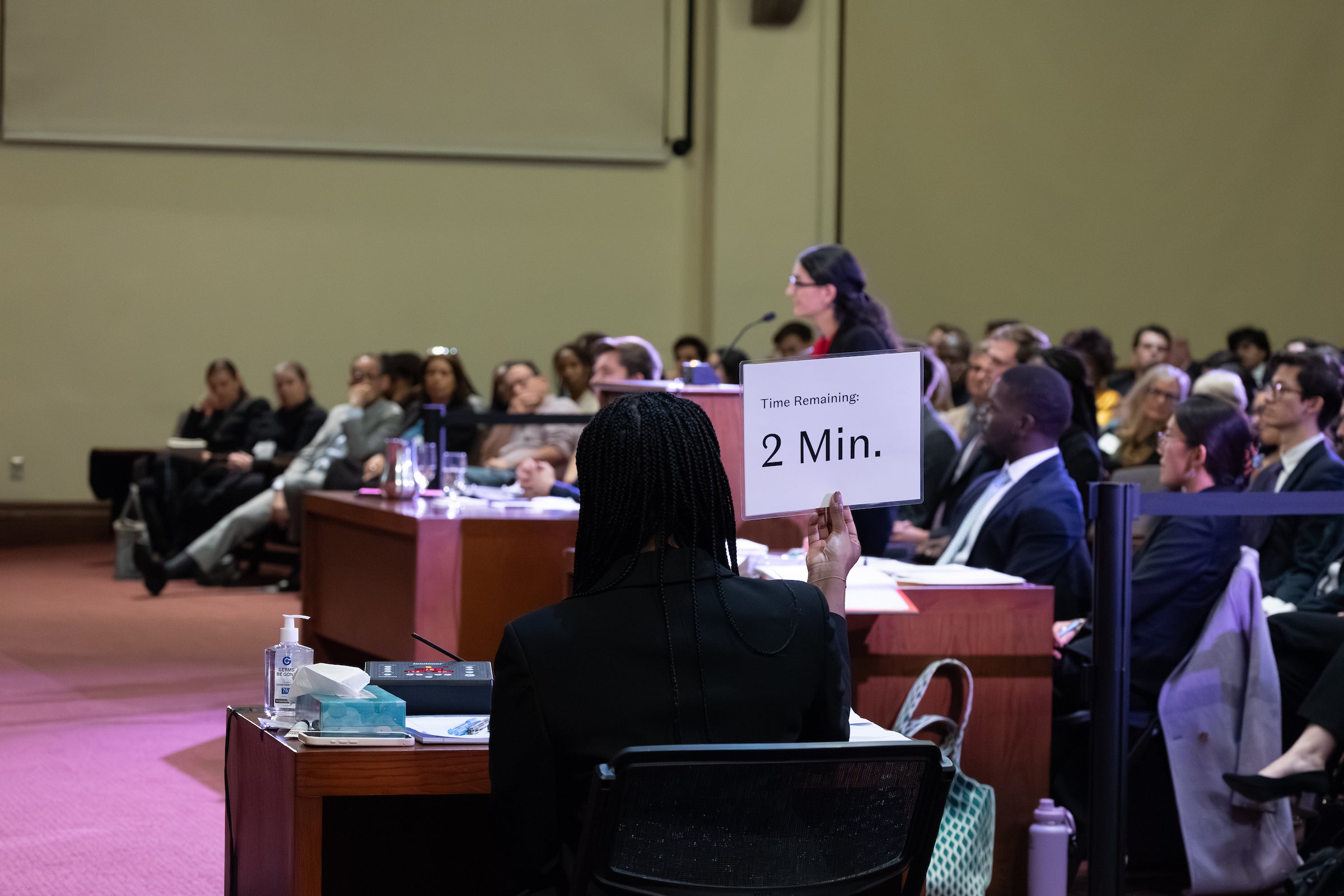 A woman holds up a sign that says 2 minutes while someone speaks at a podium.