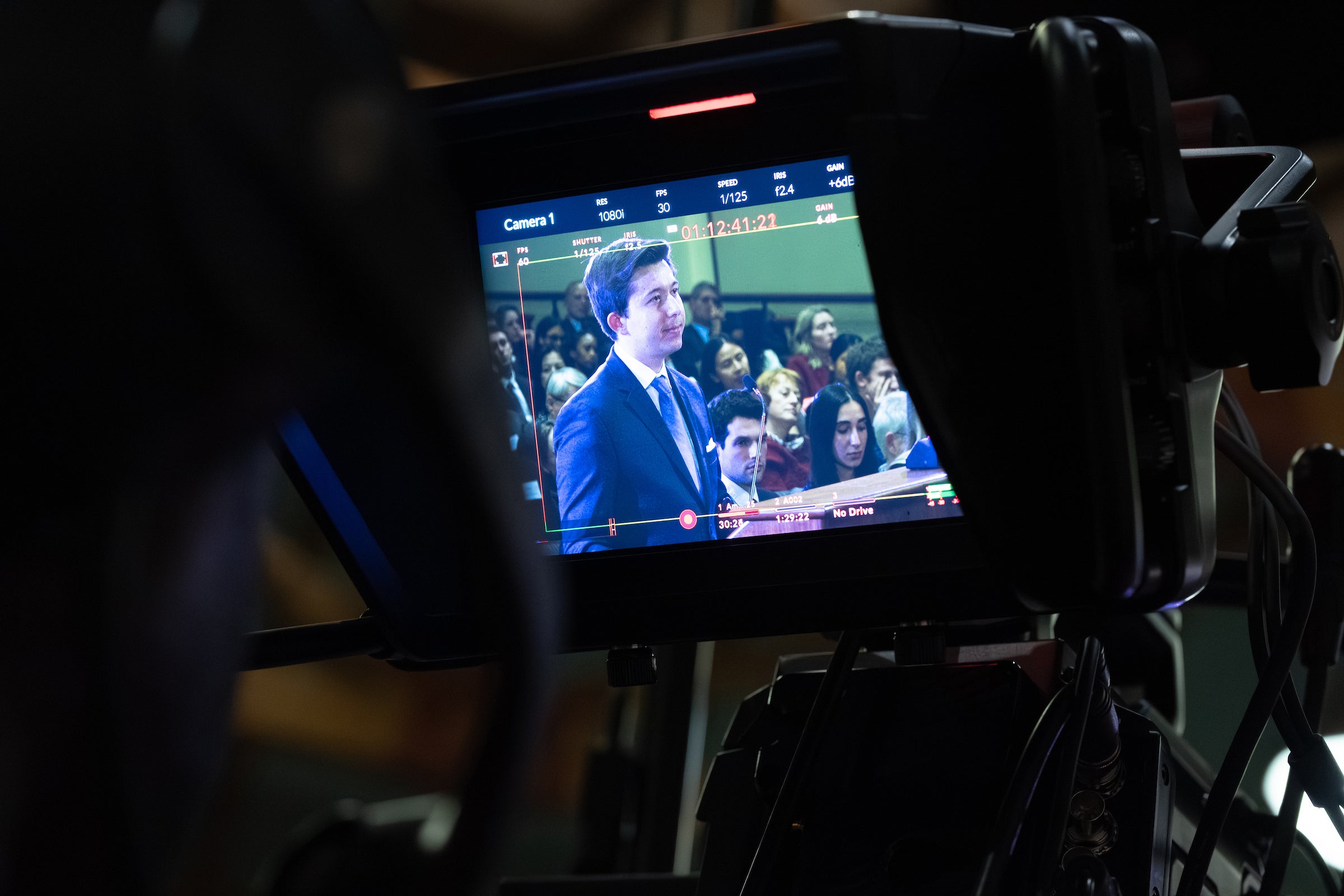 A view of a man speaking at a podium through the back of a camera.