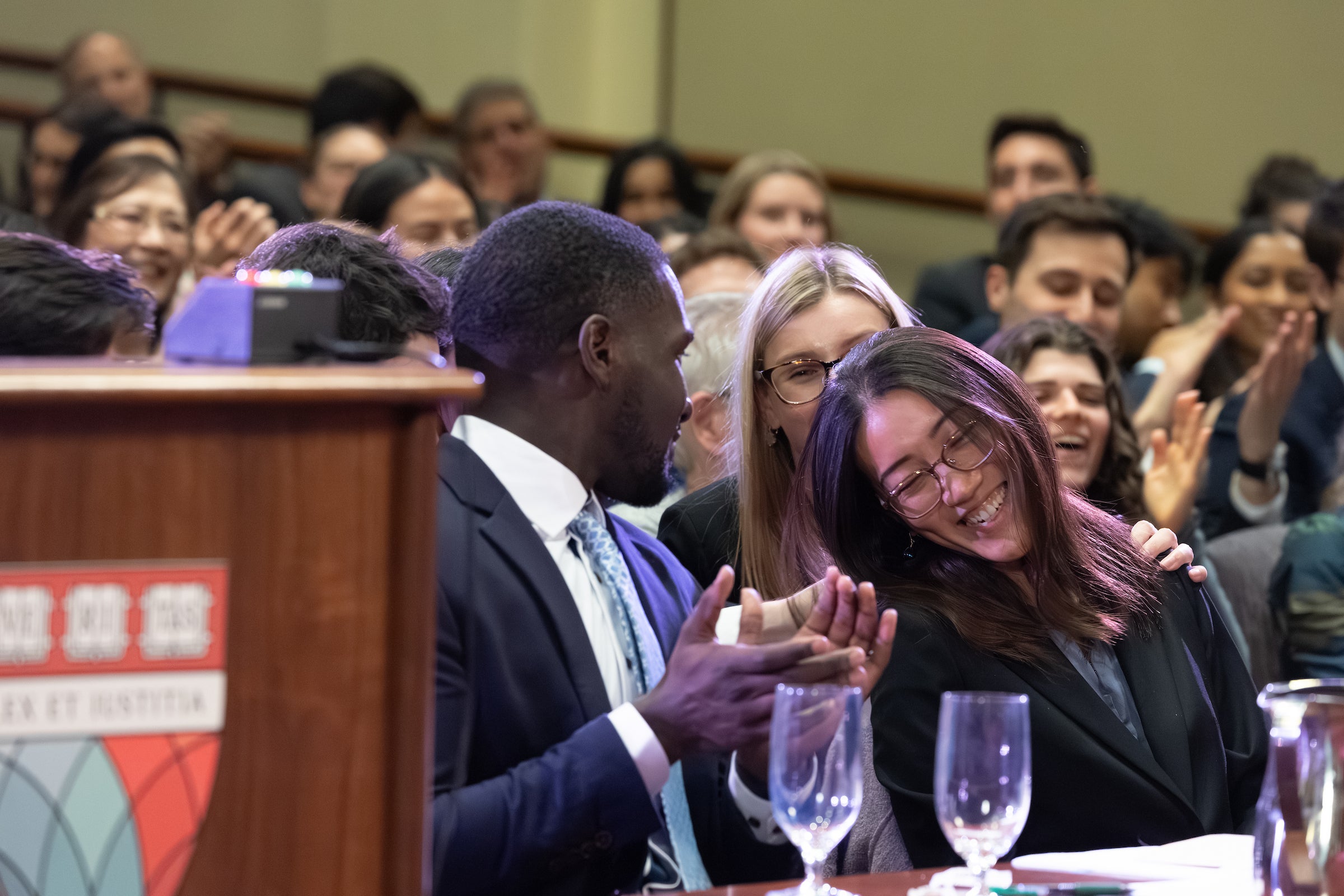 A student sitting at a table in front of an audience. responds happily to the news of a win as others clap.
