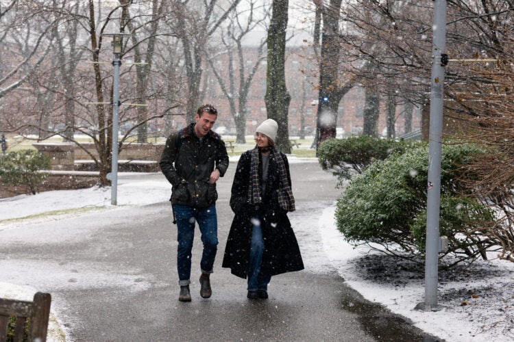 Two students walk through a campus. Snow falls through the air and is seen on the ground.