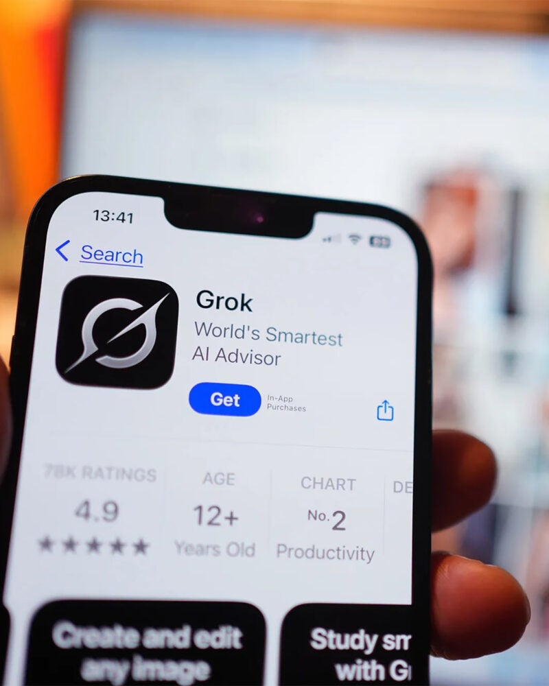 Grok app on a phone in the foreground with a laptop screen blurred in the background.