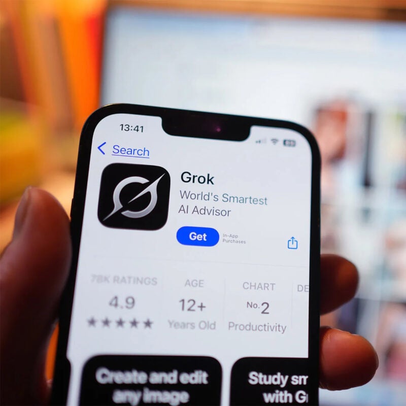 Grok app on a phone in the foreground with a laptop screen blurred in the background.