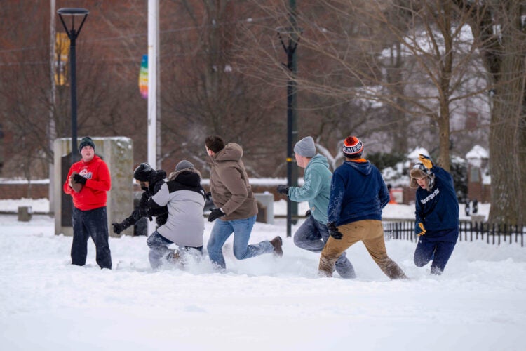 Students play flag football on a snowy field in front of bare trees.