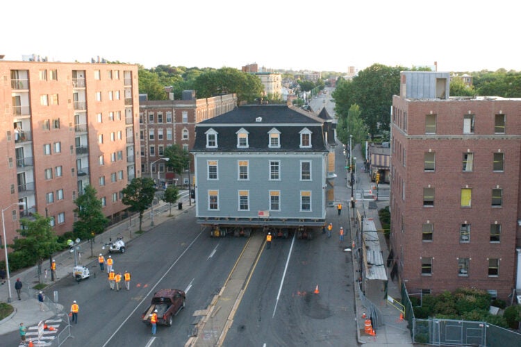 A campus building is moved down a street on hydraulic lifts. Workers in high-vis vests watch.