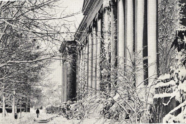 A historical black and white photograph of Langdell Hall, a stone columned building, after a snowstorm. A lone figure walks in front of the building.
