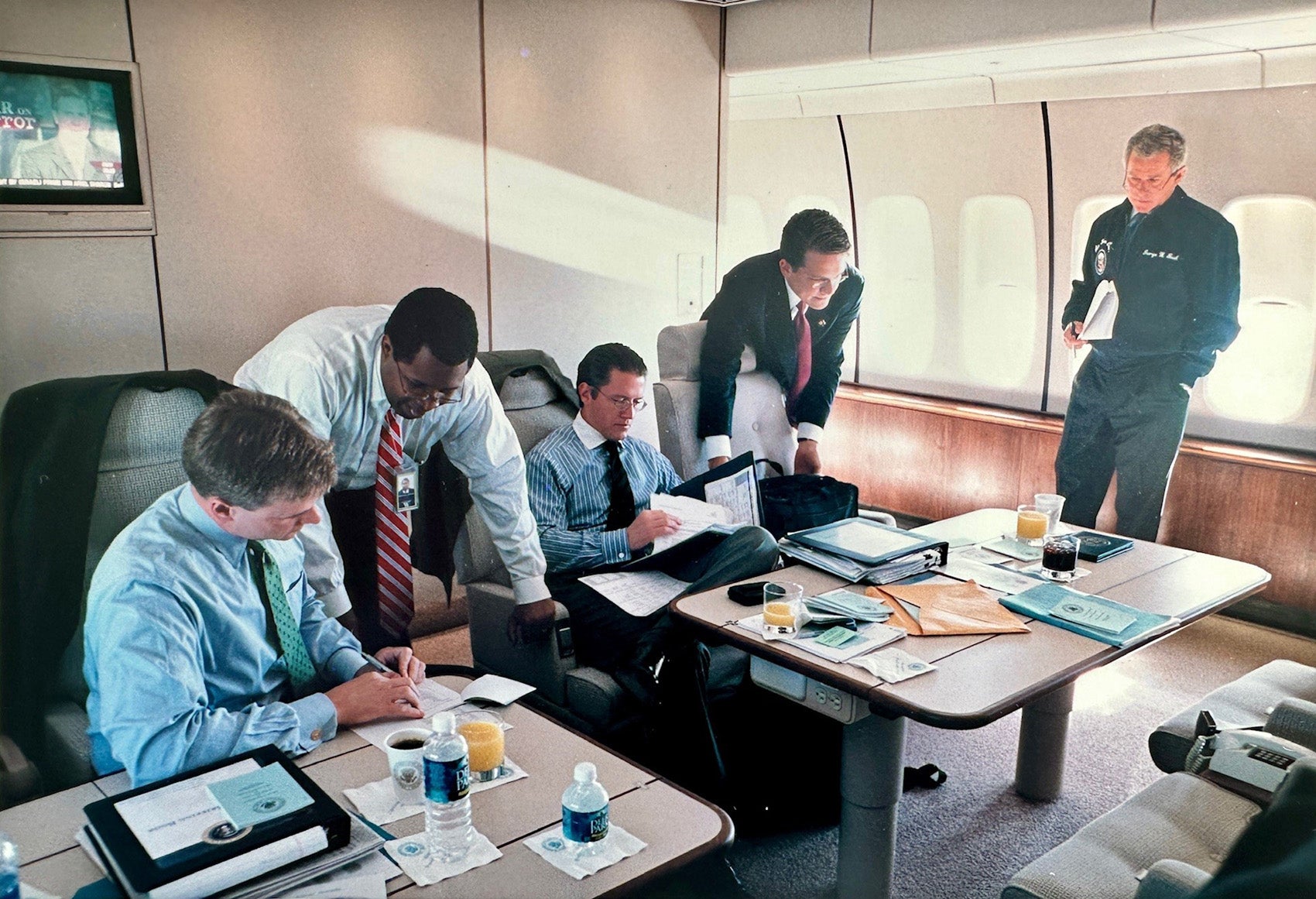 A group of men meeting at tables on an airplane while President Bush looks on.