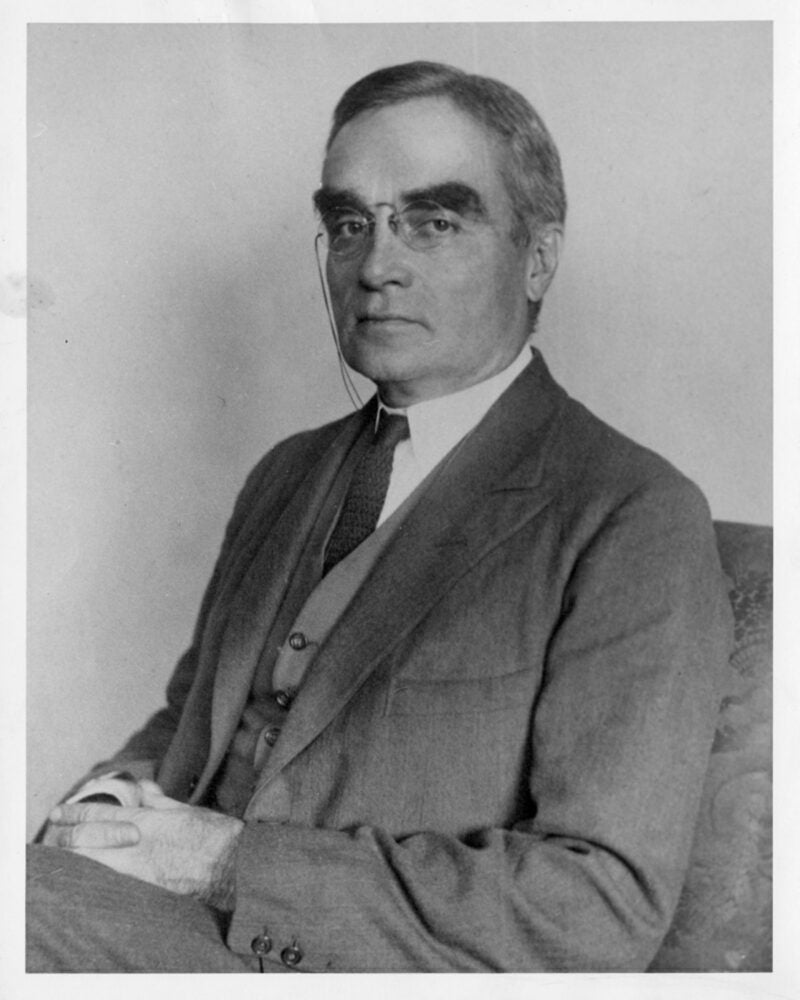 Portrait of Learned Hand sitting, hands folded in lap, wearing pince-nez glasses.