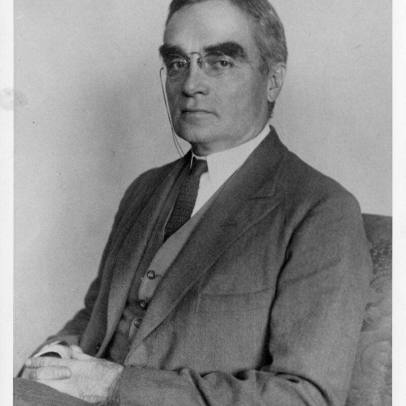 Portrait of Learned Hand sitting, hands folded in lap, wearing pince-nez glasses.