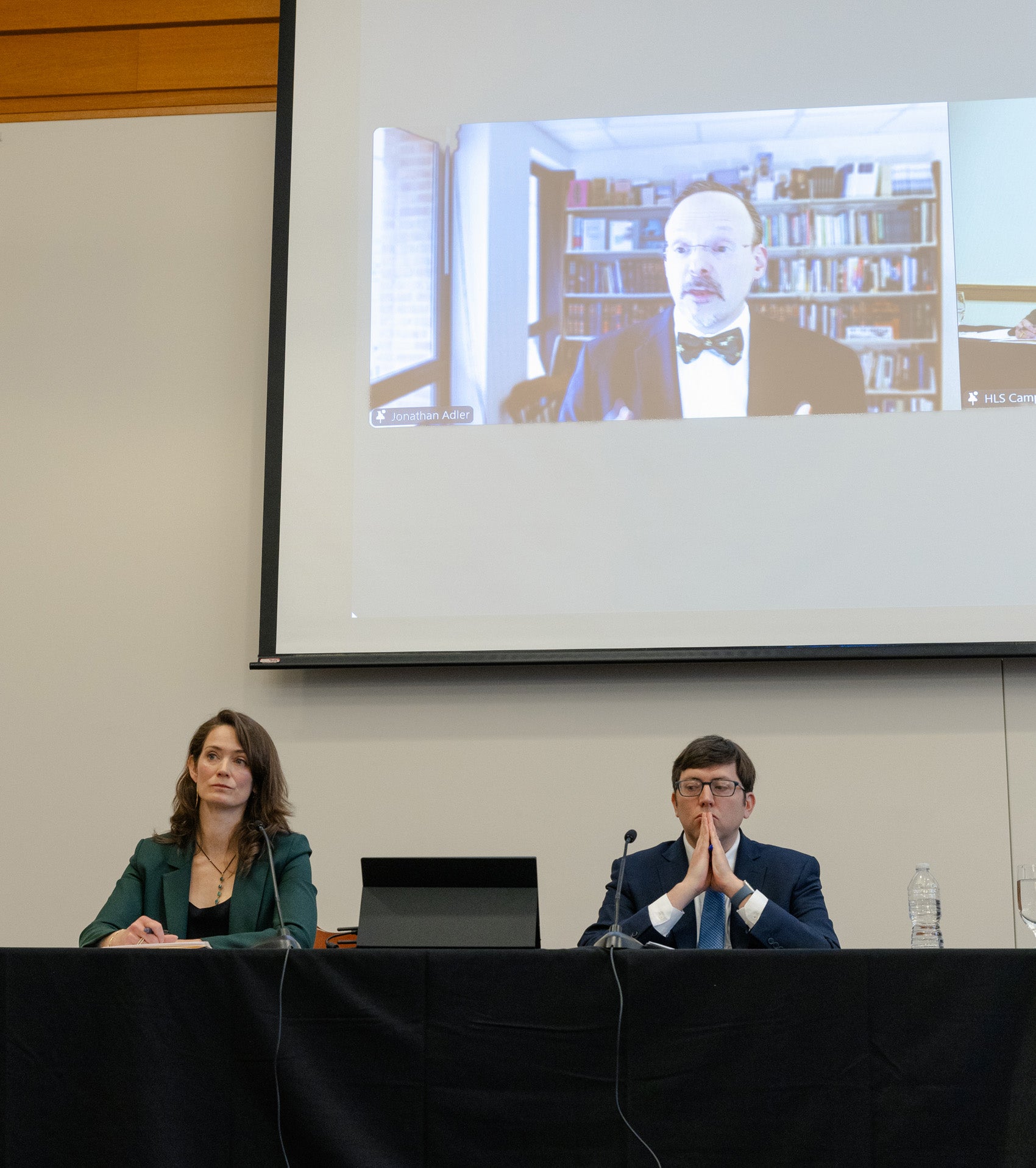 Two panelists at a table with a third on a screen in the background.