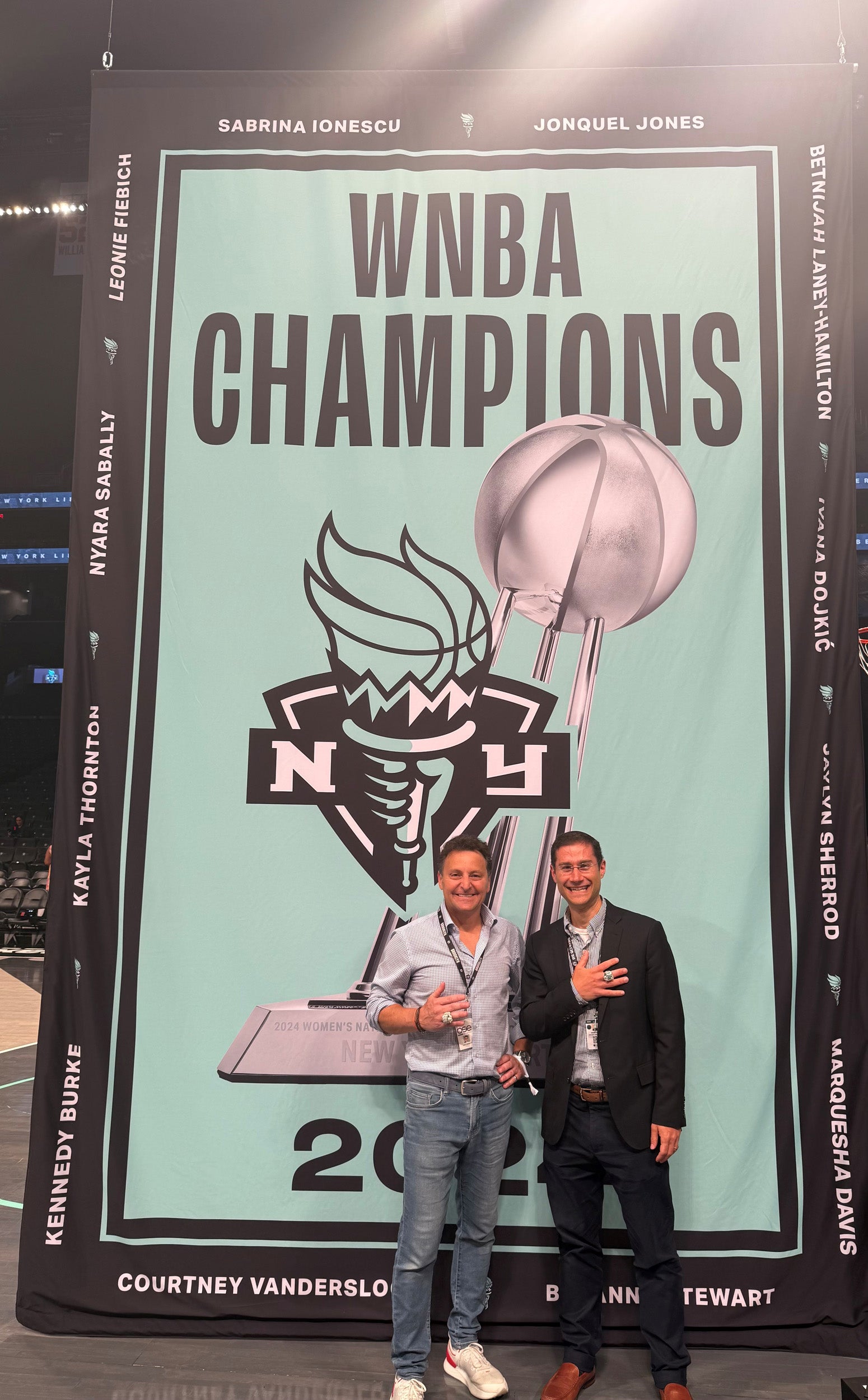Two men posing in front of a large WNBA Champions poster.