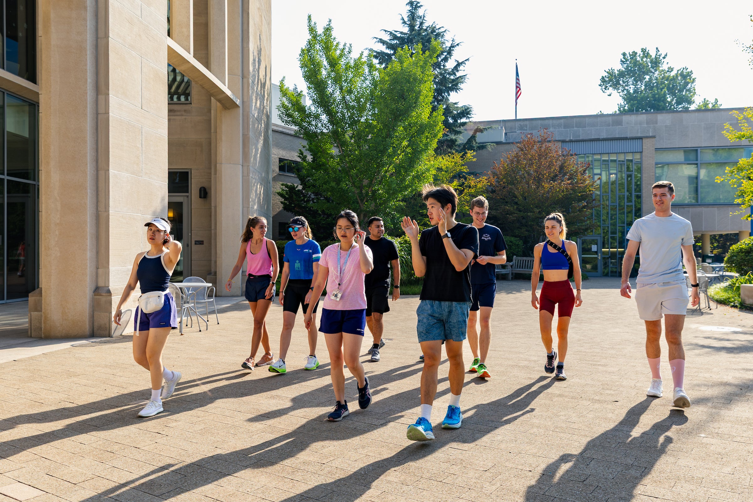 Students in running clothes walk and talk in front of a building and trees on a sunny day.