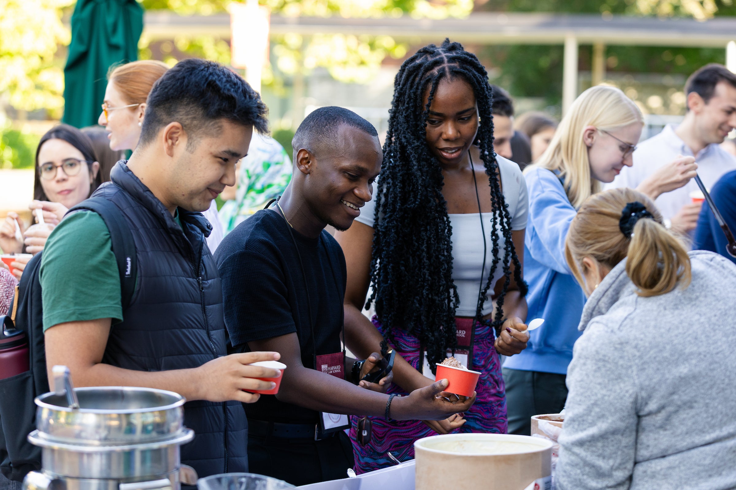 Students in a line behind a table select toppings to put on the ice cream they are holding in cups as a staff member helps them.