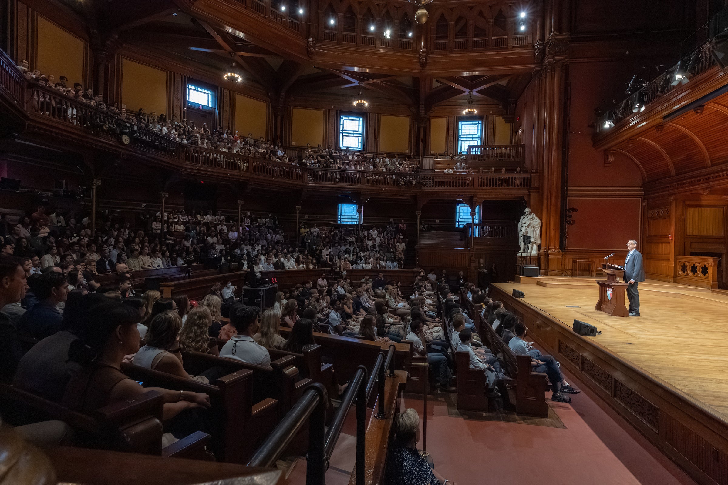 A panoramic shot of a historic theatre with dark wood walls. A large crowd of students sits in the audience to the left while a man stands on stage behind a podium to the right.