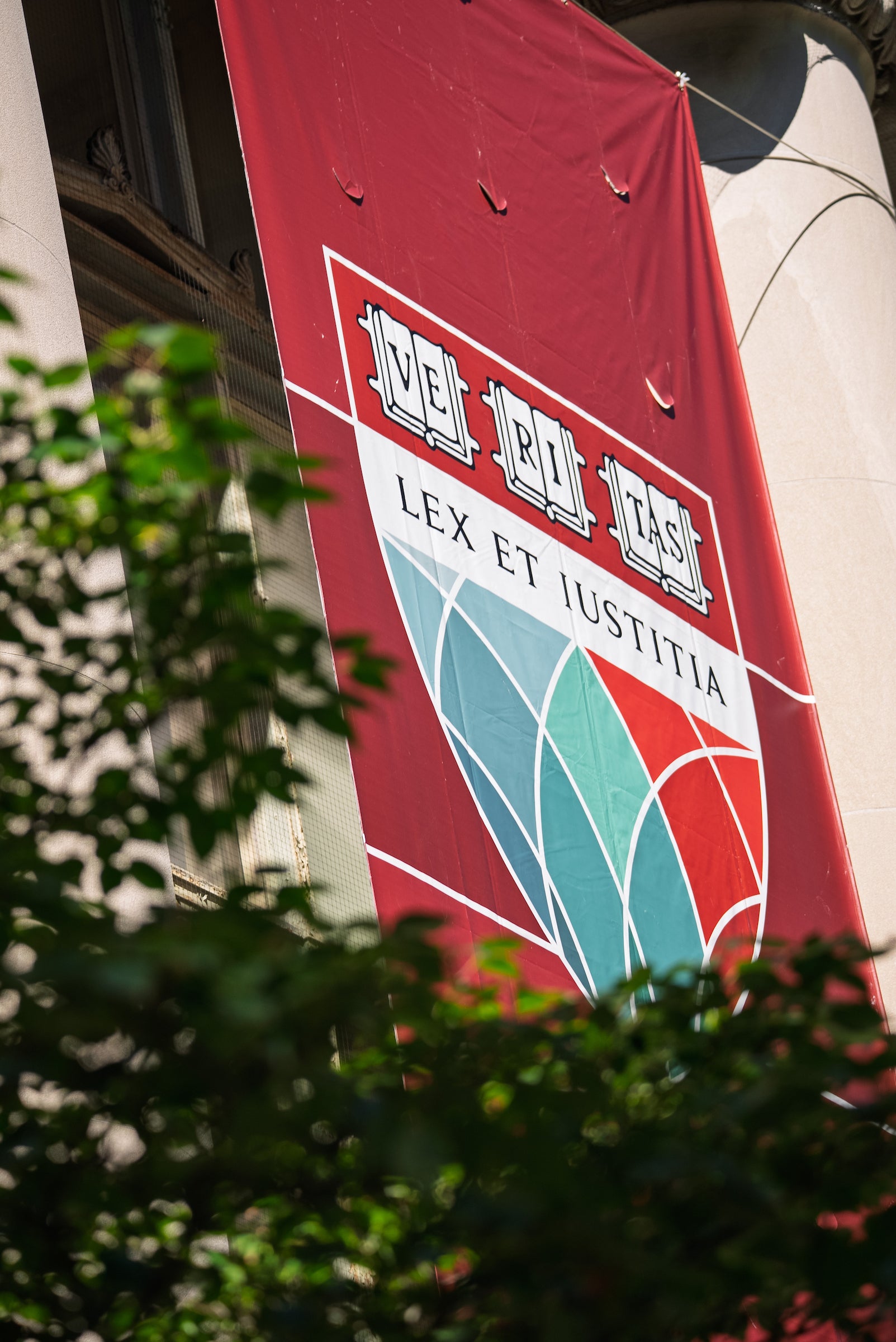 A closeup of a red Harvard Law School banner with the shield and motto, lex et iustitia, on the side of a stone building with a green tree in the corner of the frame.