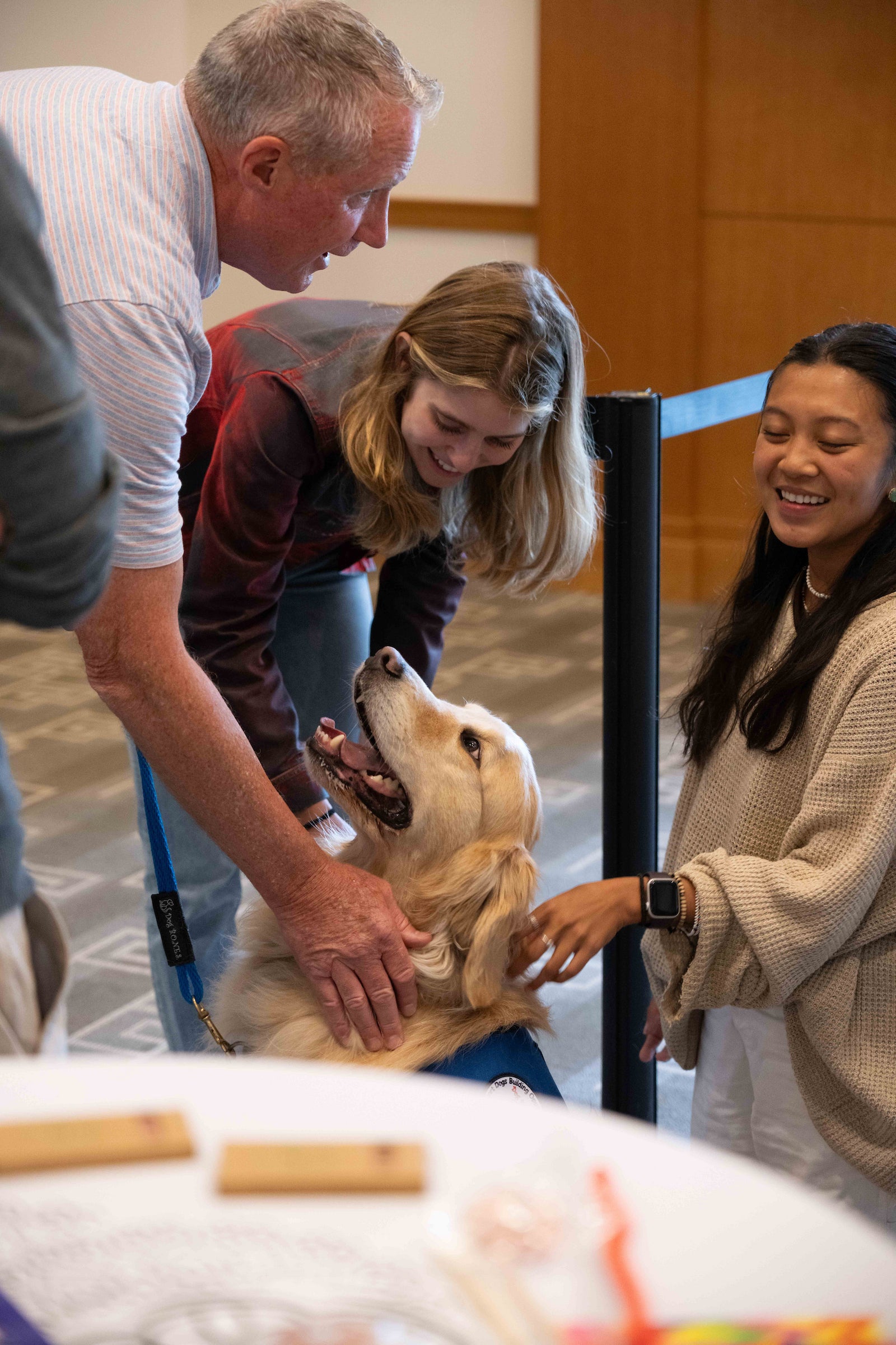 Two students, smiling, pet a dog indoors as a handler talks to them.