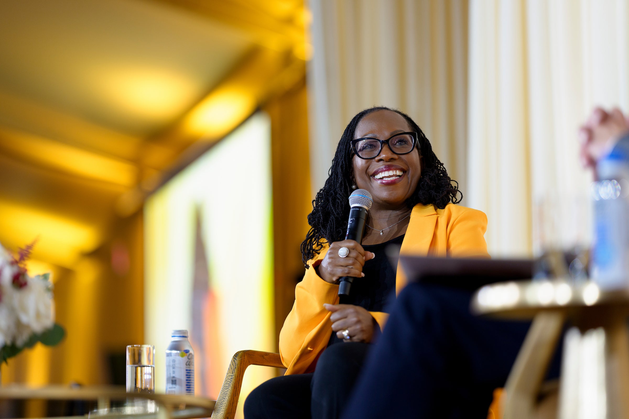 Ketanji Brown Jackson sits on a stage and speaks into a microphone in front of a yellow background.