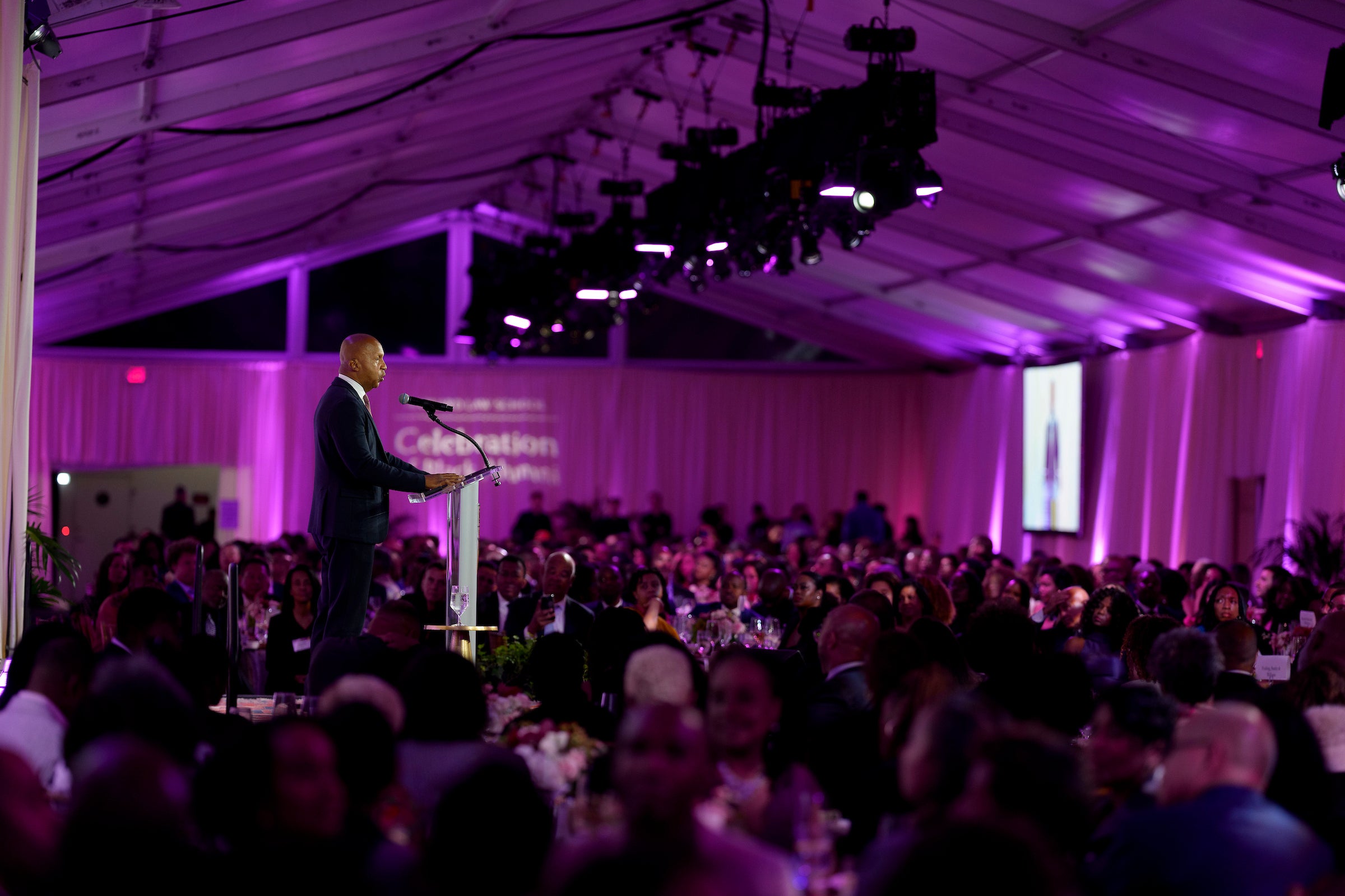 Bryan Stevenson stands on stage behind a podium to the left and addresses the audience in a tent lit with purple light.