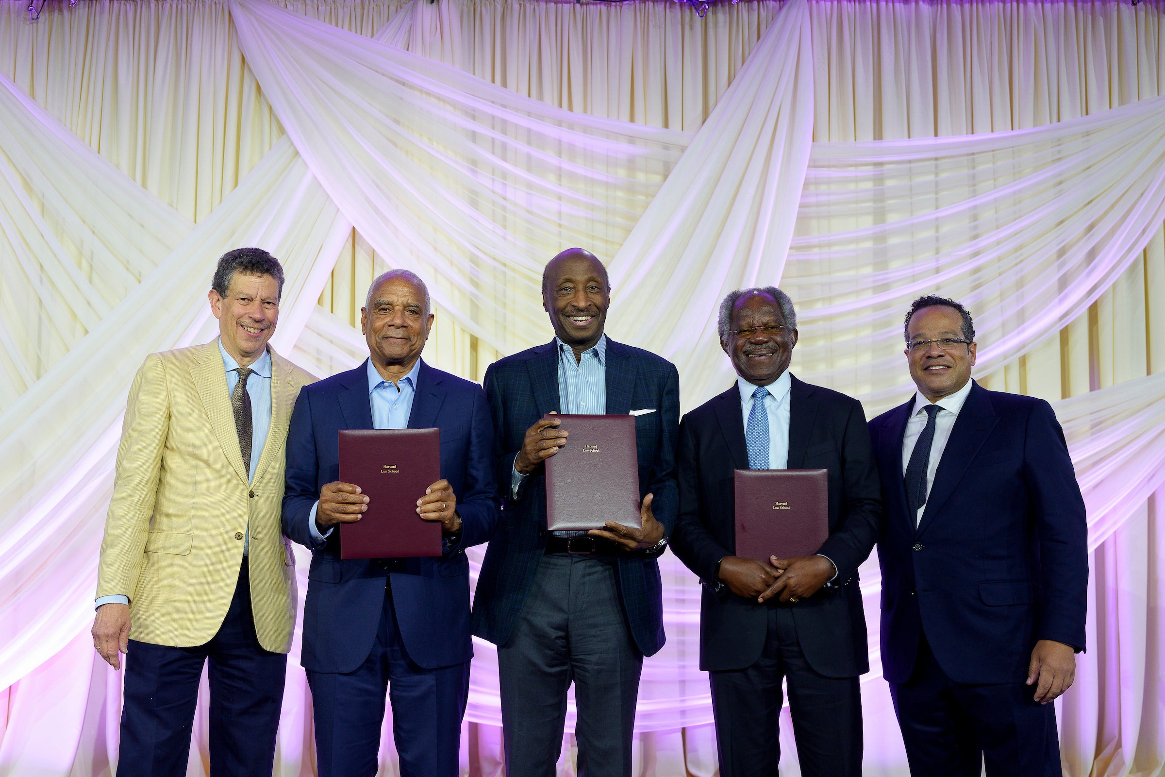 Five men in suits, three holding award plaques, pose for a photo in front of a white curtain.