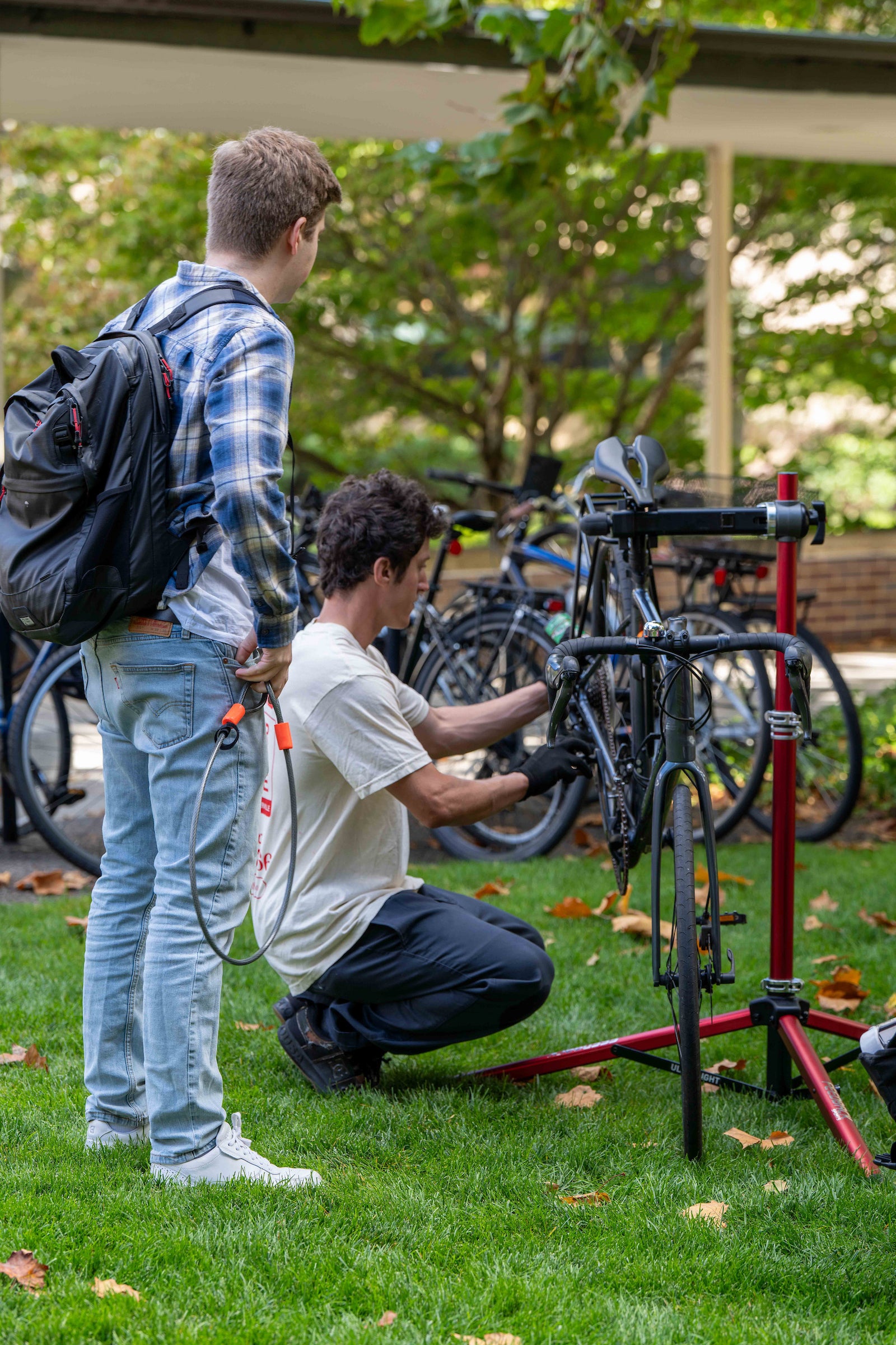 A student, standing, looks on as someone kneels down to repair his bike.
