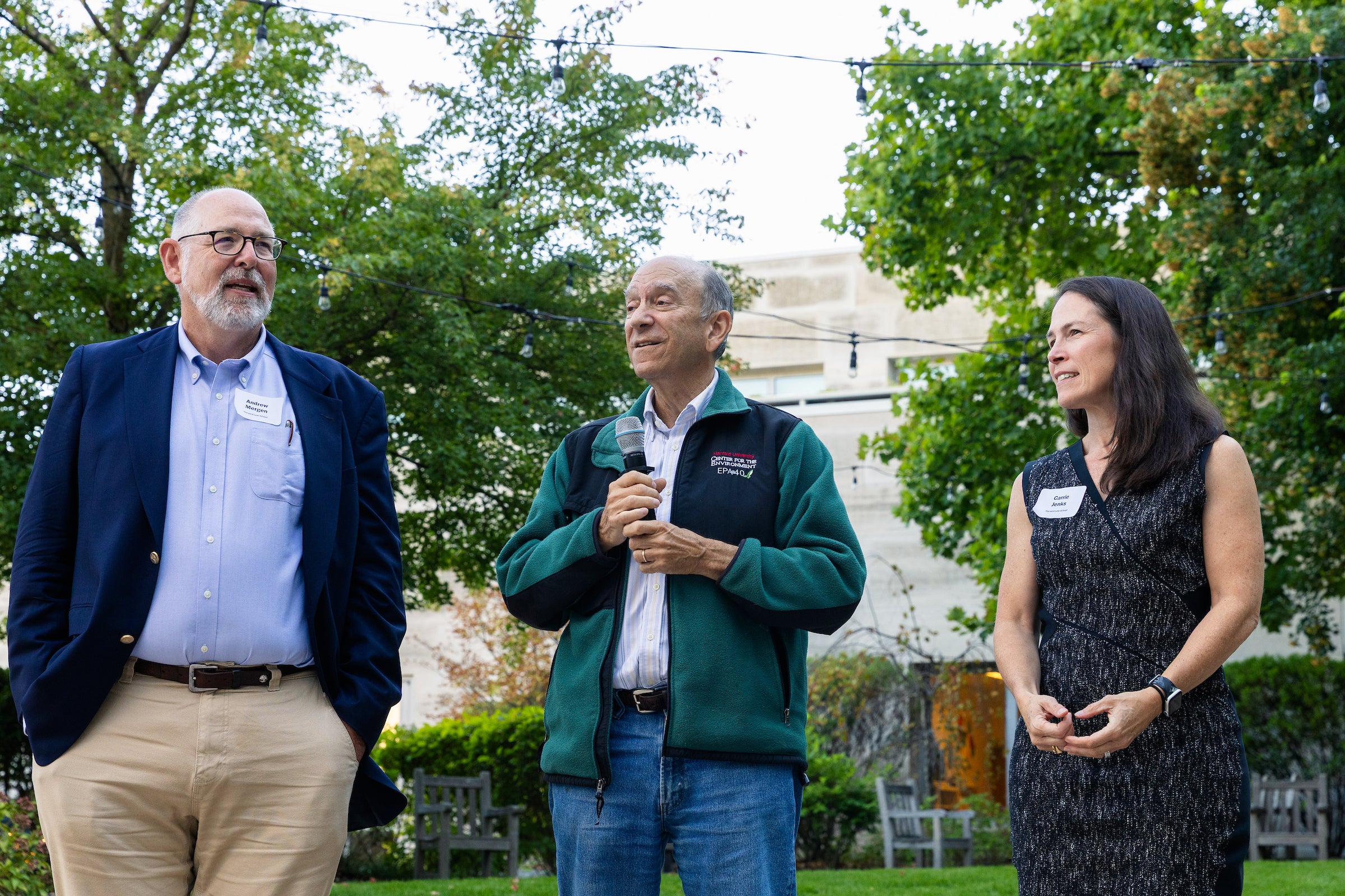 Three people stand side by side outside surrounded by trees. The person in the middle is speaking into a microphone.