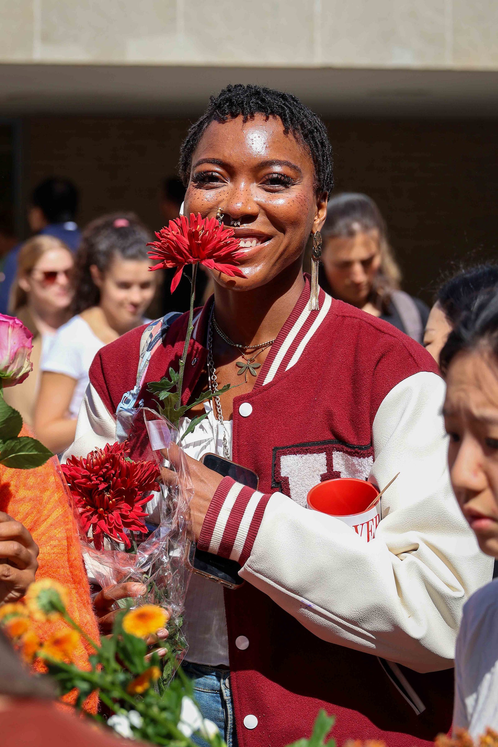 A student wearing a Harvard jacket smiles and holds up a bouquet of red flowers close to her face.
